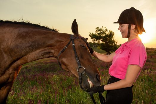 Beautiful smiling girl jockey stand next to her brown horse wearing special uniform on a sky and green field background on a sunset. Equitation sport competition and activity.