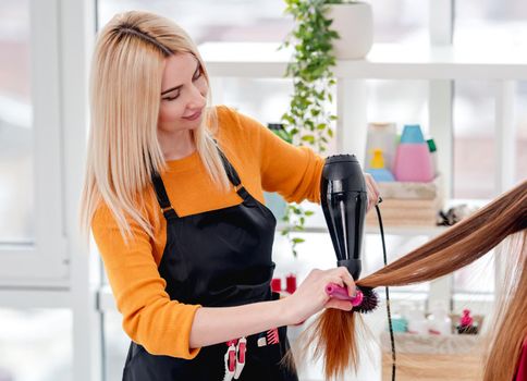 Beautiful blond hairdresser care about long hair of client with hairdryer