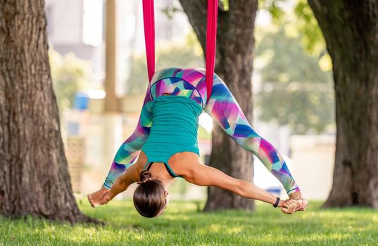 Girl during fly yoga practice in hammock stretching her body at nature. Young woman doing aero fitness gymnastic exercising outdoors