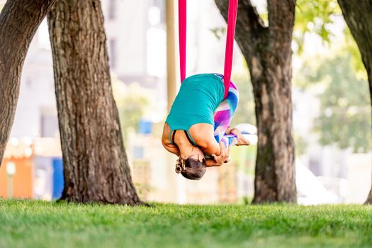 Sport girl practicing fly yoga in hammock at nature and staying in child pose in the air. Young woman doing aero fitness gymnastic stretching outdoors