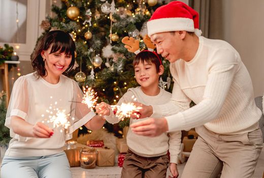 Happy family in santa hats with sparklers sitting under decorated christmas tree at home