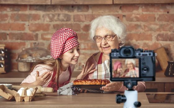 Little granddaughter with her senior granny enjoying the smell of hot homemade fruit pie. Blogger profession. Shooting with a camera for a blog