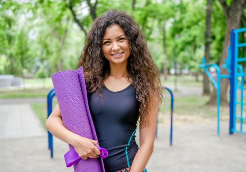 Beautiful girl with yoga mat standing at the stadium outdoors. Young woman exercising outside