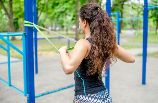 Sport girl with elastic rubber band doing arm workout outdoors. Young woman exercising with equipment