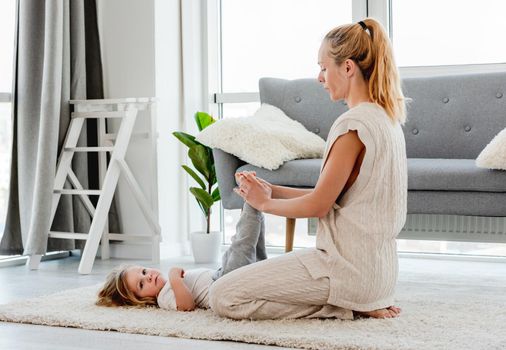Young blond mother making exercises for her child son legs on the floor at home