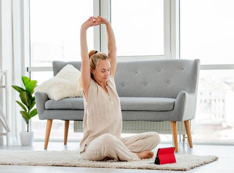 Blond attractive girl stretch her back during online yoga class in tablet. Home workouts in covid time