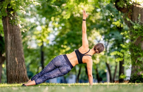 Girl doing yoga at nature and standing in side plank pose with arm up. Young woman during stretching portrait from back in summertime outdoors
