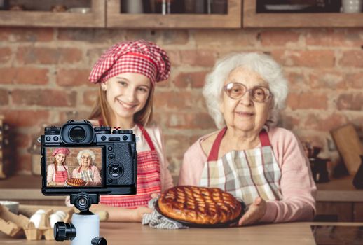 Young little granddaughter with her granny holding a tray and showing homemade fruit pie that they baked together. Blogger profession. Shooting with a camera for a blog