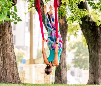 Sport girl practicing fly yoga in hammock at nature and keeping balance in the air with body down and legs up. Young woman doing aero fitness gymnastic stretching outdoors