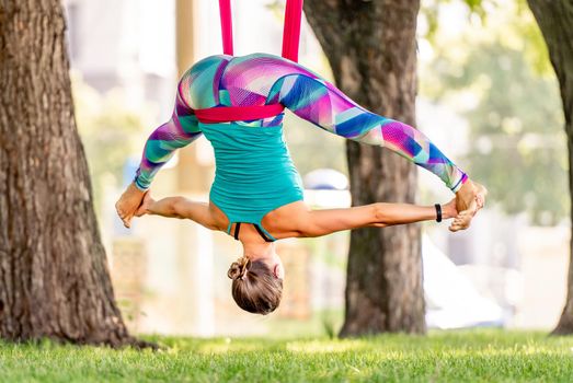 Sport girl practicing fly yoga in hammock at nature and doing leg split twine in the air. Young woman doing aero gymnastics outdoors
