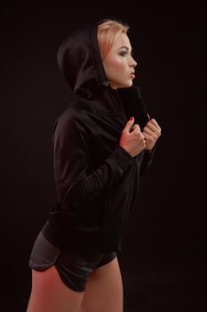 Beautiful seductive sporty girl with the slim sexy figure in the black hoodie, sport underwear and little black shorts is posing in the studio. olor filters.