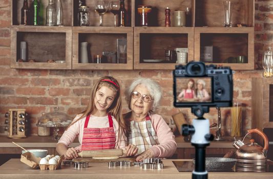 Eldery granny with her lovely granddaughter roll out the dough using the rolling pin and looking at camera. Blogger profession. Shooting with a camera for a blog