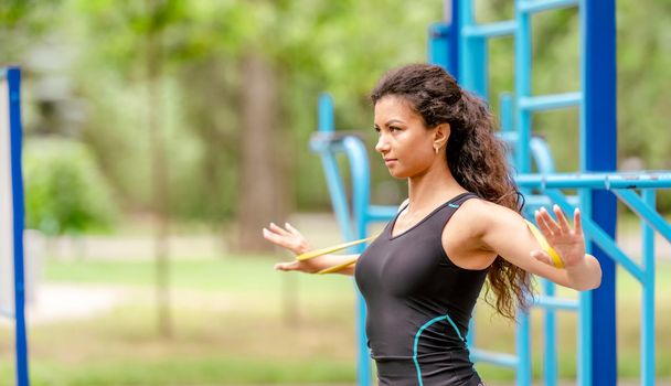 Sport girl with elastic rubber band doing back and arm workout outdoors. Young woman exercising with fitness equipment at stadium