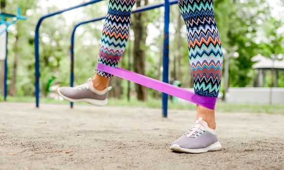 Girl with elastic rubber band doing leg workout. Young woman exercising outdoors at the stadium