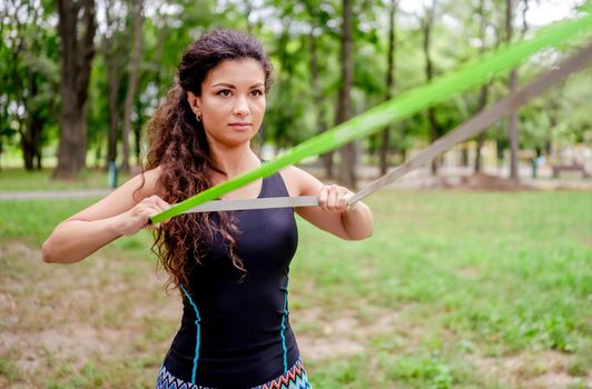 Sport girl with elastic rubber band doing arm workout outdoors. Young woman exercising with equipment at summertime