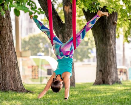 Girl during fly yoga practice in hammock stretching her body at nature. Young woman doing aero fitness gymnastic exercising outdoors at summer