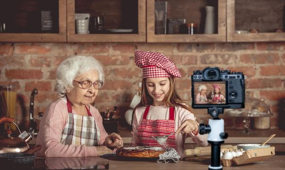 Granny helps granddaughter sprinkling sugar powder with sieve on homemade cake. Blogger profession. Shooting with a camera for a blog