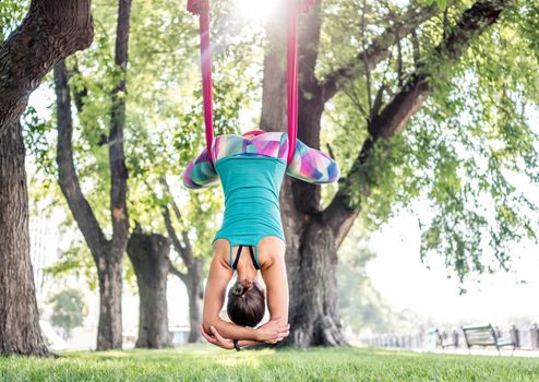 Sport girl practicing fly yoga in hammock at nature and stretching keeping her body down. Active woman during aero gymnastics outdoors