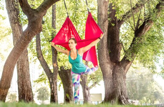 Sport girl practicing fly yoga in hammock at nature and stretching her body standing on one leg. Active woman during aero gymnastics outdoors