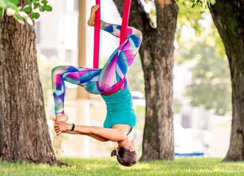 Sport girl practicing fly yoga in hammock at nature and stretching her legs in the air. Young woman doing aero gymnastics and keeping balance
