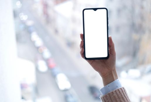 Woman holding phone with blank screen in hand on street view from window background