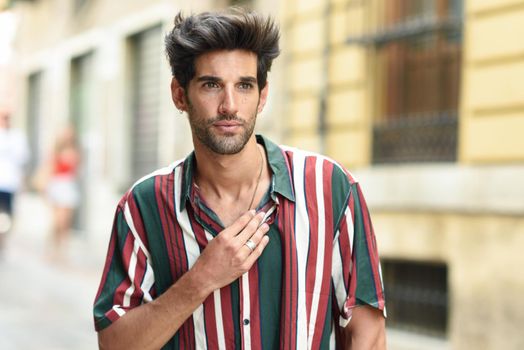 Handsome young man with dark hair and modern hairstyle wearing casual clothes in urban background.