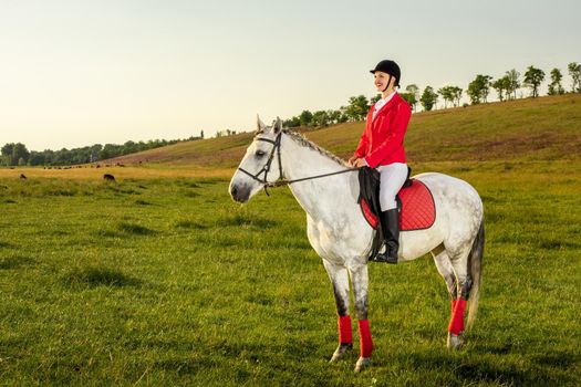 Young woman rider, wearing red redingote and white breeches, with her horse in evening sunset light. Outdoor photography in lifestyle mood