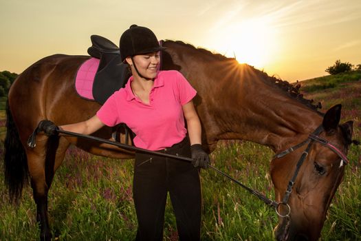 Beautiful smiling girl jockey stand next to her brown horse wearing special uniform on a sky and green field background on a sunset. Equitation sport competition and activity.