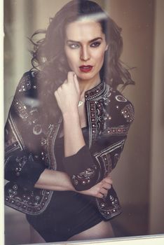 Young brunette woman looking through the window. Attractive girl, model of fashion, wearing modern jacket and black lingerie. Female with long wavy hairstyle.