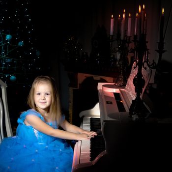 Beautiful little girl is playing on a white piano in a dark room by candlelight. The concept of Christmas and New Year, family happiness.