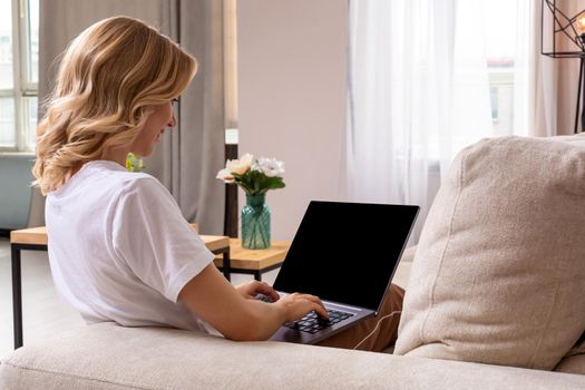 Woman on sofa working on laptop with mockup blank screen. Empty copy space on monitor for advertisement, language learning ad, online shopping website, social media site. Black screen.