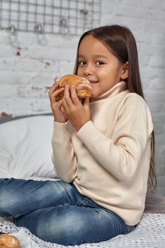 Pretty little girl 6 years old in a white sweater and jeans. Child in the room with a bed, eating croissant and drinking tea or cocoa, good mood.