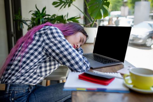 Young woman lying on her arms on the table in cafe in front of laptop with cup of coffee, sleepy. Rear shot of female student learning online via laptop before her lectures.