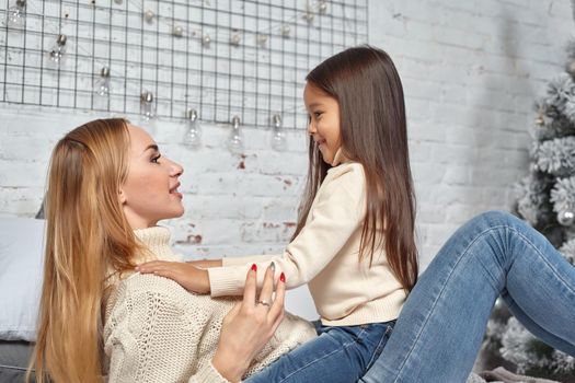 Beautiful young woman and her charming little daughter in white sweaters and jeans lying on the bed at home, are hugging.