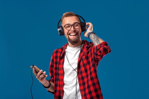 Happy young man listening to music with headphones. Handsome smiling guy in checkered shirt dancing with headphones. Isolated on blue background. Concept of people emotions