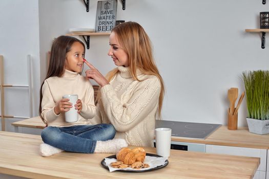 Shot of cheerful mother and daughter sit together at kitchen table, drink hot tea in morning, have pleasant friendly talk between each other. Curious girl asks something in mum during coffee break.