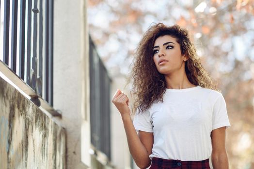 Beautiful young arabic woman with black curly hairstyle. Arab girl in casual clothes in the street. Happy female wearing white t-shirt and checked pants.