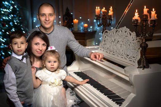 A happy family with their young children in a dark room by candlelight, near a large white piano. The concept of holidays and happy childhood.