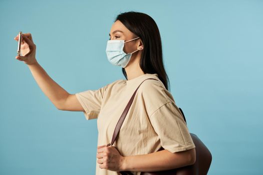 Side view of woman holding smartphone while making selfie in studio with backpack, isolated on blue background. Copy space. Quarantine, epidemic concept