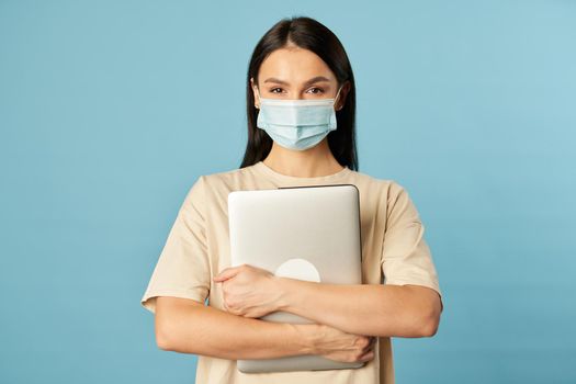 Waist up of young woman wearing face mask and holding laptop while looking at camera, isolated on blue background. Copy space. Quarantine, epidemic concept