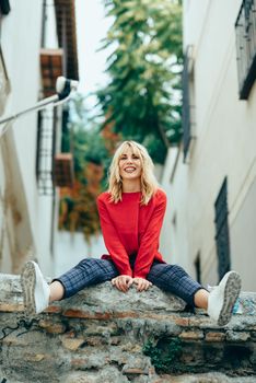 Happy young blond woman sitting on urban background. Smiling blonde girl with red shirt enjoying life outdoors.