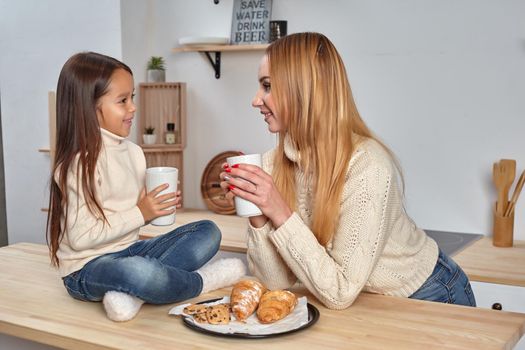 Shot of cheerful mother and daughter sit together at kitchen table, drink hot tea in morning, have pleasant friendly talk between each other. Curious girl asks something in mum during coffee break.