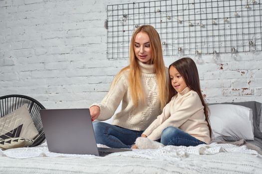 Beautiful young mother and her cute daughter in white sweaters and jeans lying on the bed at home, laughing and looking in laptop. Family time