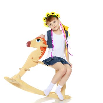 A beautiful little blonde girl in a summer short dress, and a wreath of yellow flowers on her head.The girl is rocking on a wooden horse.