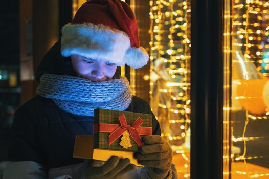 Boy in Santas hat with gifts box near illuminated shop window. Xmas presents holidays, or shopping on New Year or Christmas concept