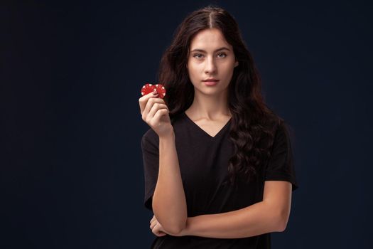 Curly hair brunette wearing a blackish t-shirt is posing with red gambling chips in her folded hands and looking at the camera. Poker concept on a black background. Casino.
