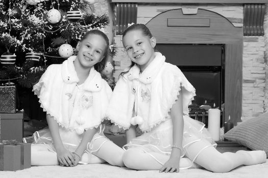 Cute little twin girls, sitting on the floor near the Christmas tree and electric fireplace on which candles are burning. In the hands of the girls boxes with gifts.Black-and-white photo. Retro style.