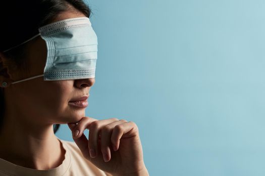 Close up of lady in a beige t-shirt posing with a protective mask on her eyes on a blue background. Copy space. Quarantine, epidemic concept