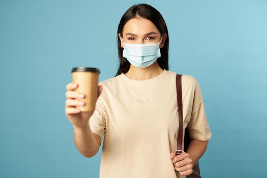 Happy young female wearing face mask posing with hot drink and backpack while posing in studio against blue background. Copy space. Quarantine, epidemic concept
