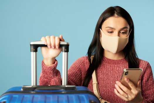 Pretty lady in protective mask from virus and looking on the mobile phone while expecting flight on blue background. Copy space. Concept of travel, coronavirus